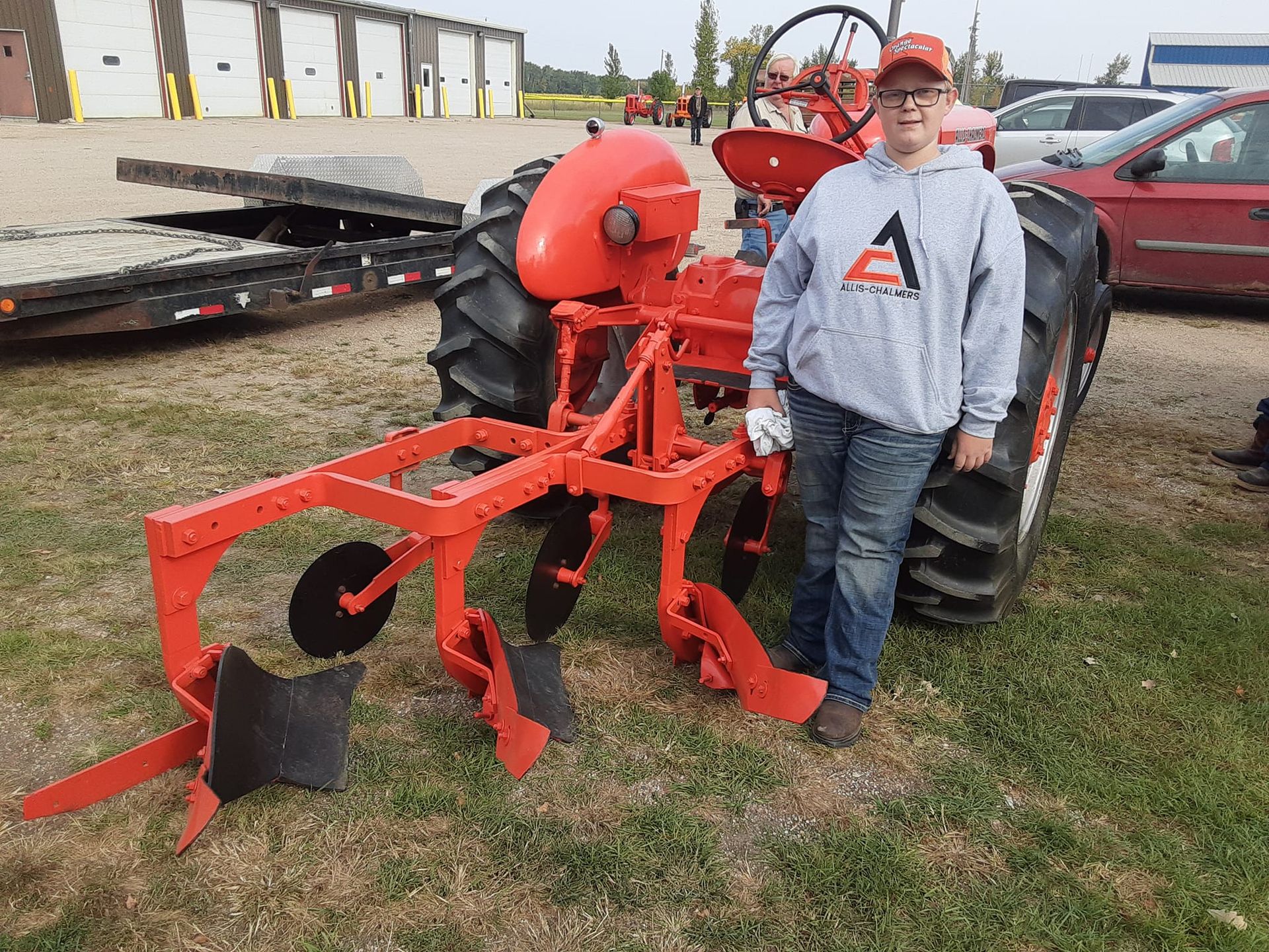 Person stands by an orange tractor and plow on grass field.