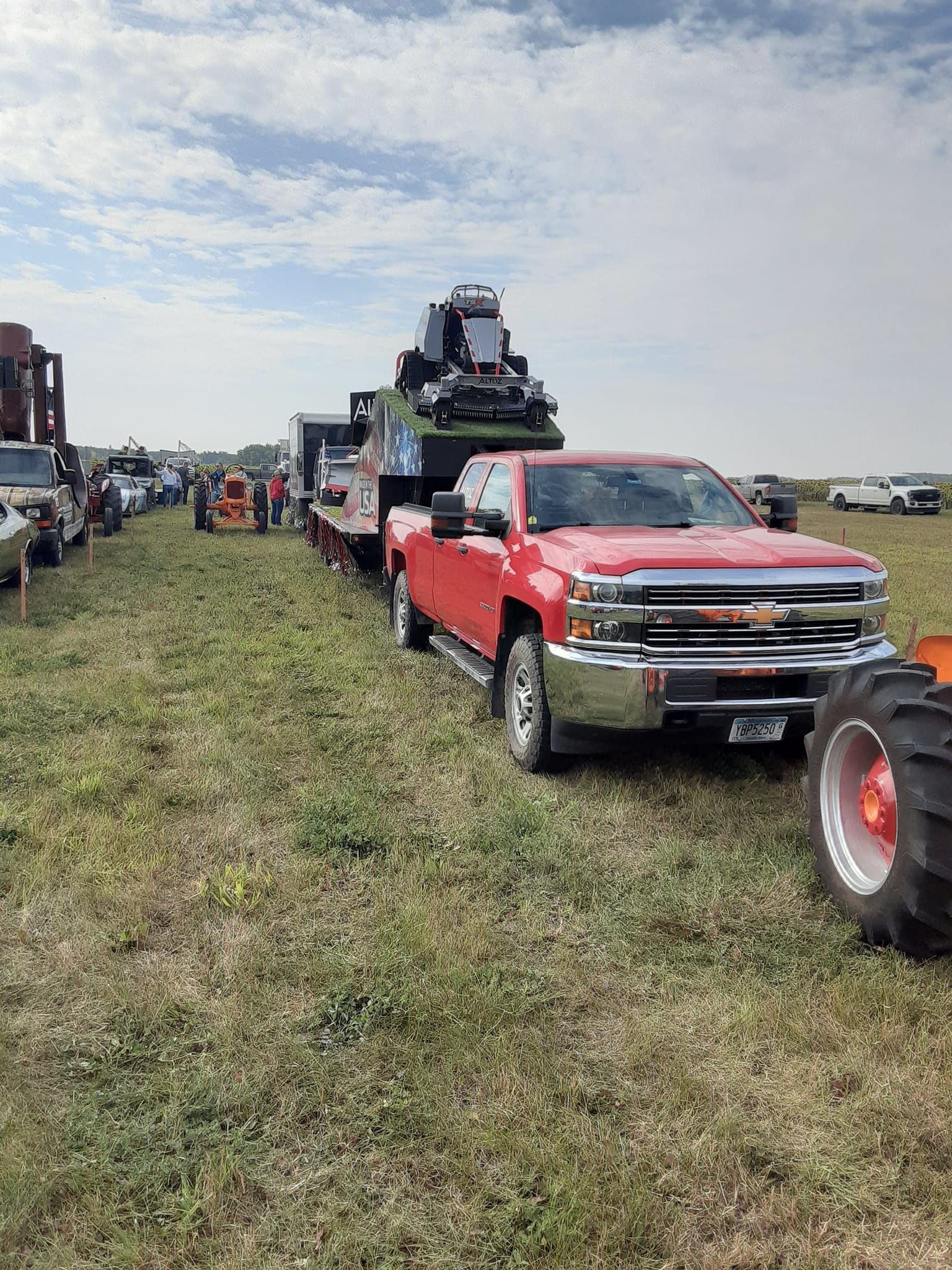 A bright red pickup truck hauls a flatbed trailer with a mechanical loader across a grassy field at an outdoor event.
