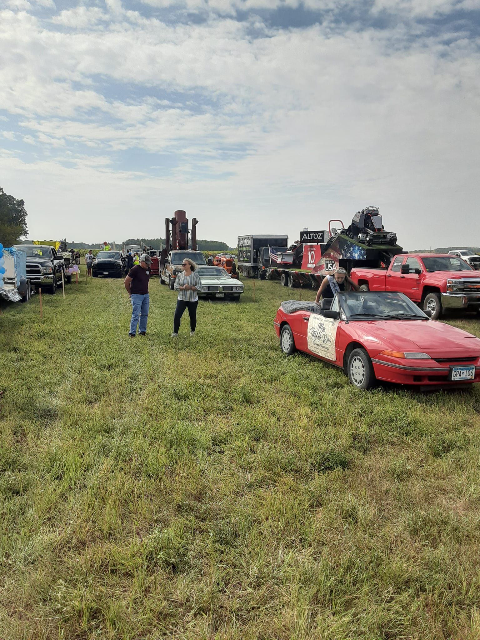 People and cars in a grassy field. Red car in foreground, others in the background under a cloudy sky.