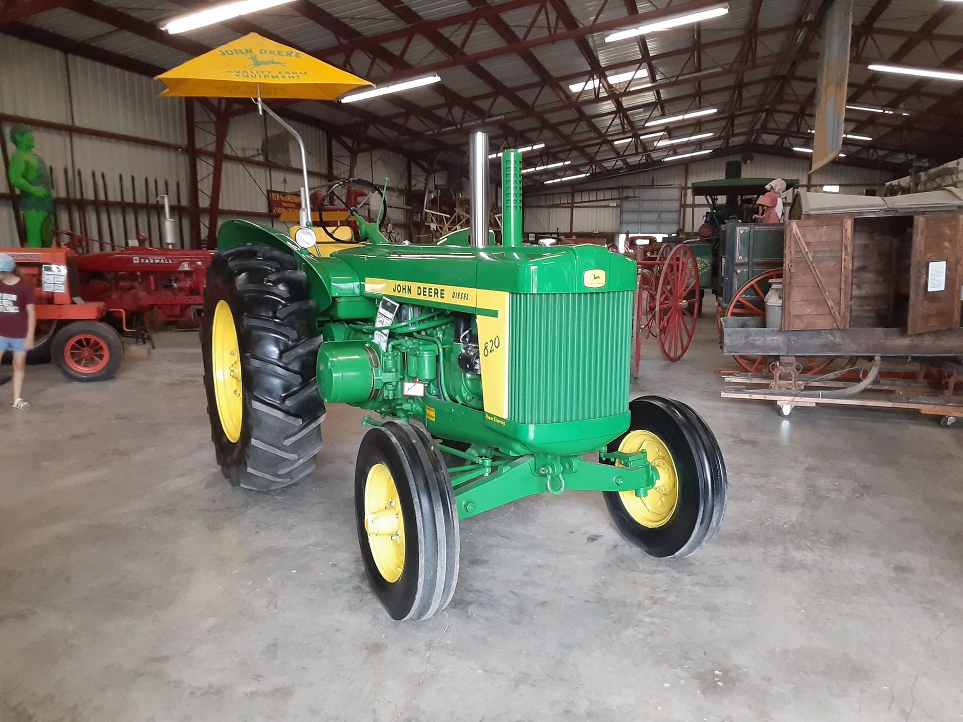 A bright green John Deere tractor with a yellow canopy parked inside a large, open-air equipment museum.