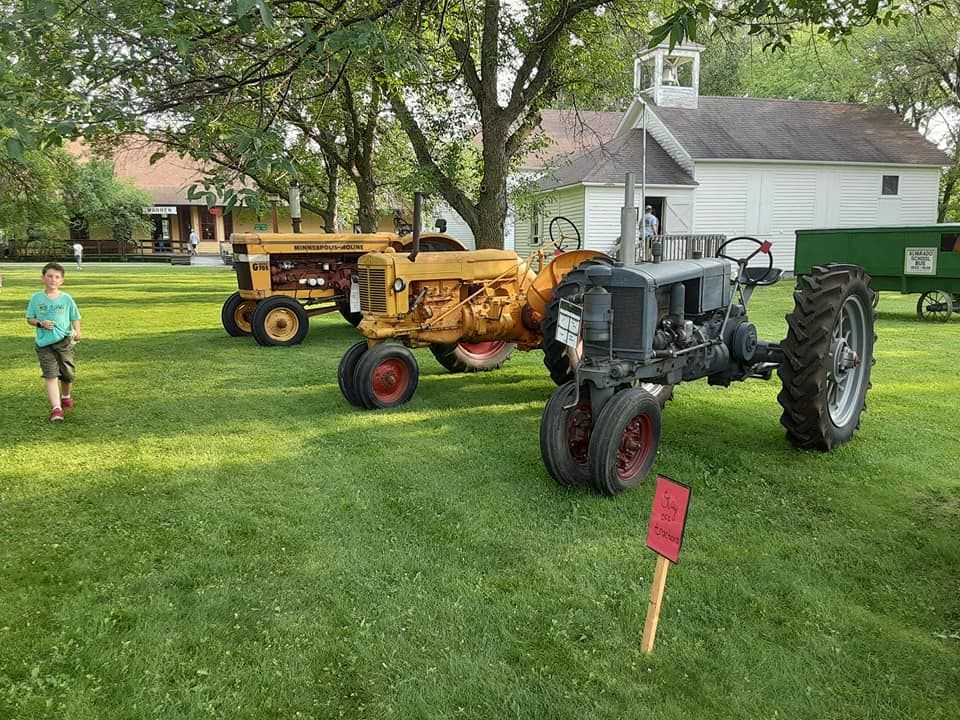 Three vintage tractors on green grass with a small schoolhouse in the background. A boy stands nearby.