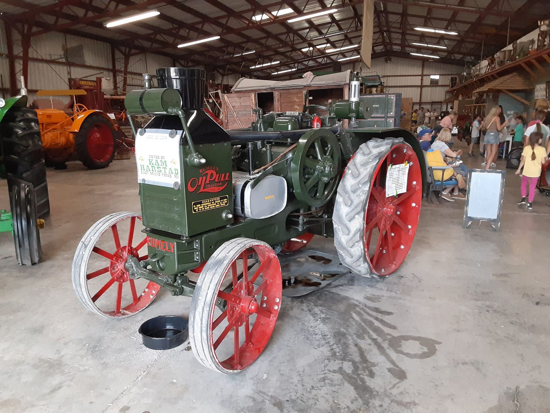 Green antique tractor with red wheels on display in a large building with onlookers.