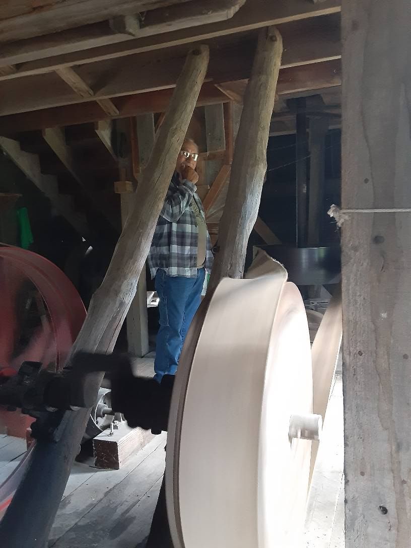 Man standing near large machinery inside a wooden building; he wears a plaid shirt and jeans.