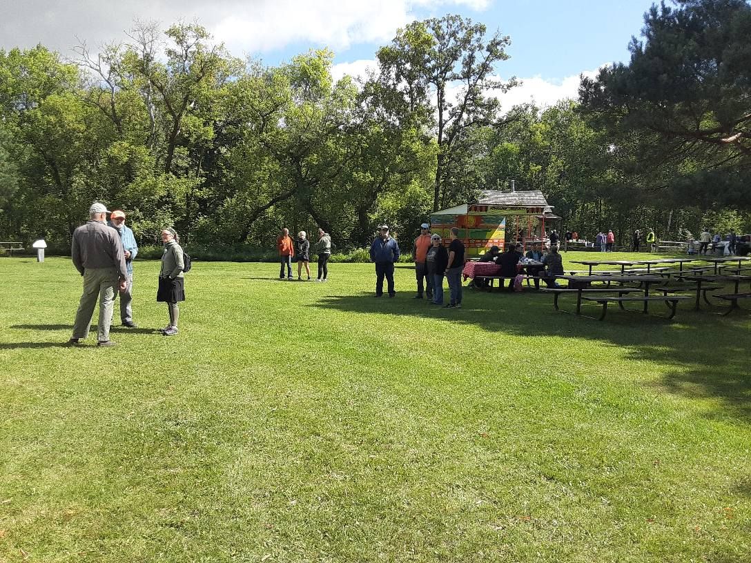 People gather in a sunny park with a food truck, tables, and trees.