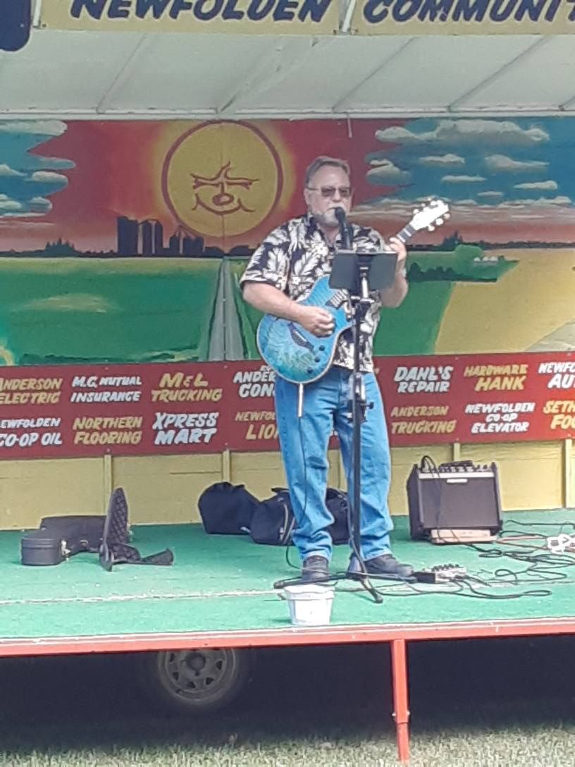 Man playing a blue guitar and singing on a stage, possibly outdoors.