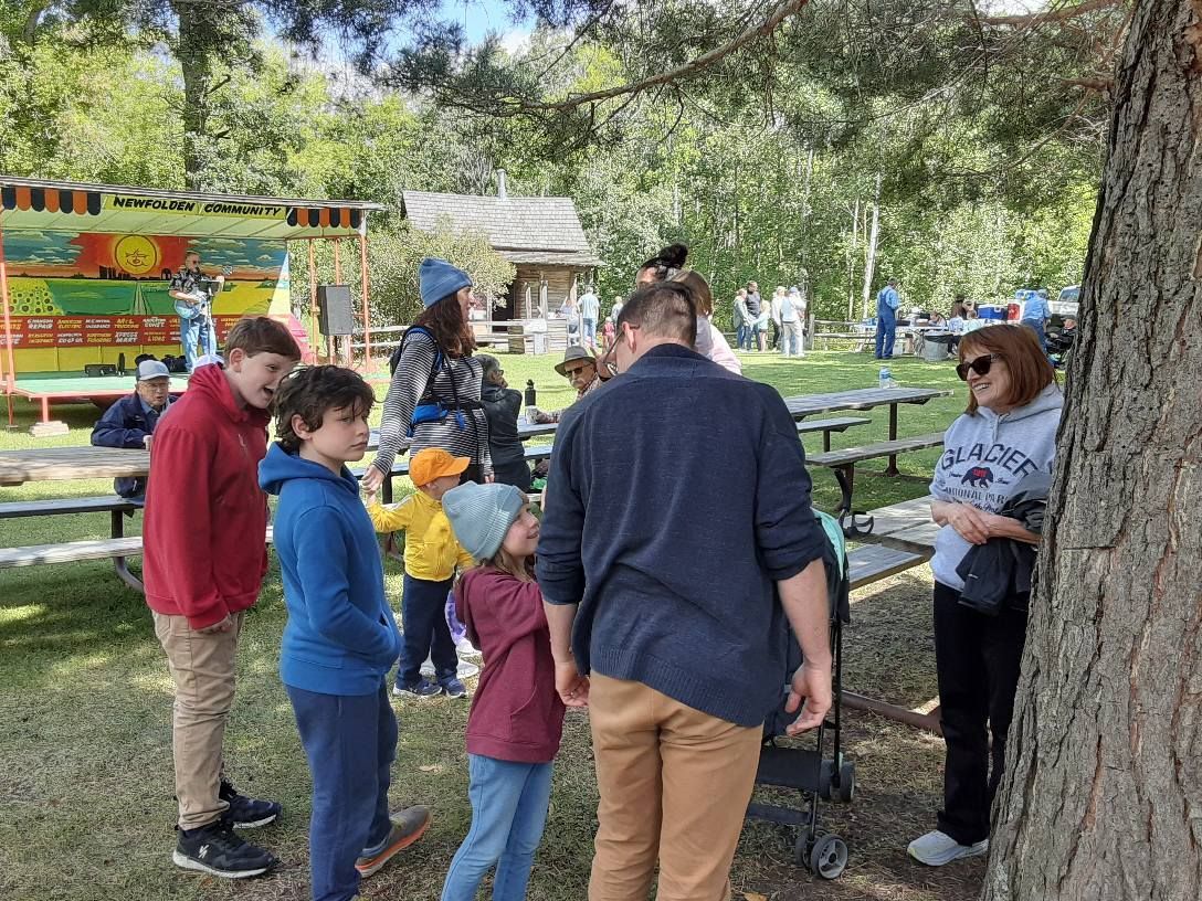 Group of people gathered outdoors, near a wooden structure and picnic tables. Sunlight and trees are visible.