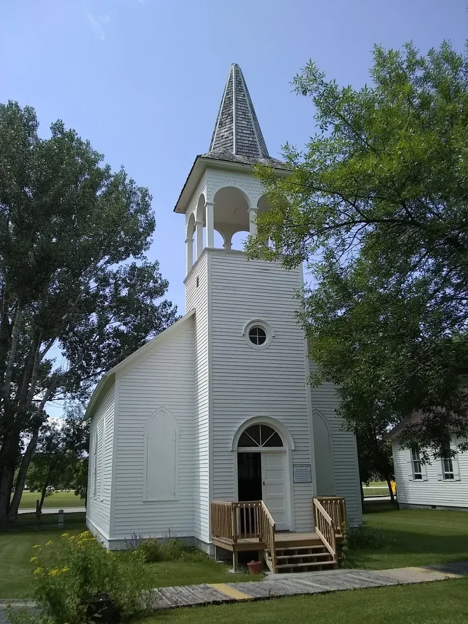 A white wooden church with a tall steeple, an open front door, and a wooden entrance ramp, set in a grassy landscape.