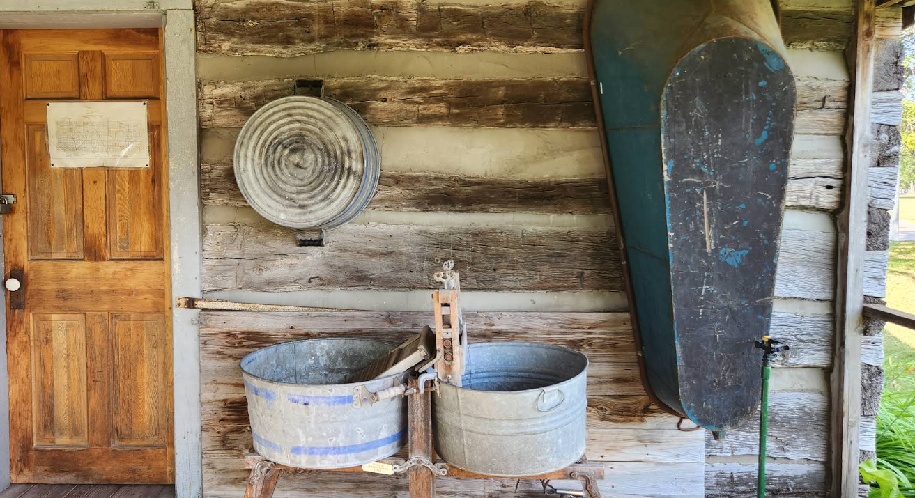 Wooden door, rustic wash station with metal tubs, coiled tub, and water tank on log cabin exterior.