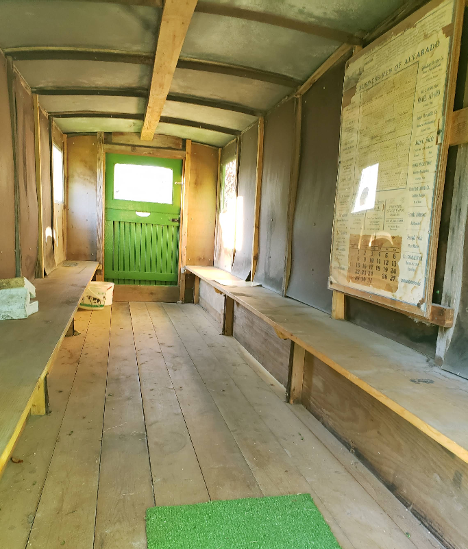 Interior of a wooden cabin with benches, a green door, and a document board. Natural light streams in.