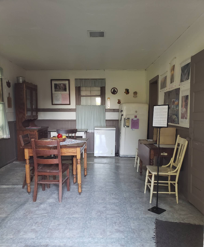 Kitchen with wooden furniture, including table and chairs, and a refrigerator. Wall posters and a window visible.