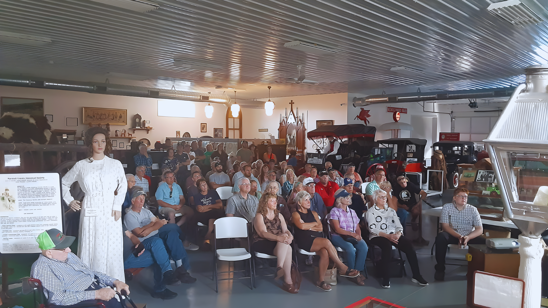 An audience seated in chairs in a large, industrial-style venue listens to a presentation given by a person in white.