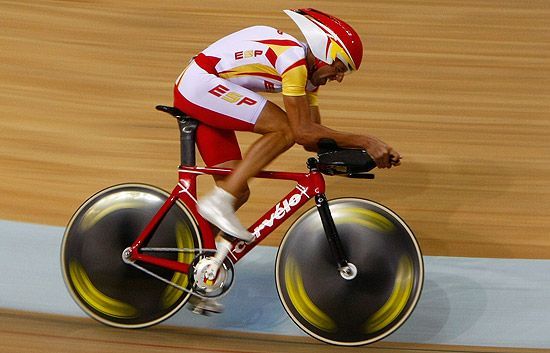 Ciclista con uniforme español en una bicicleta Cervelo roja, circulando sobre una pista de madera con ruedas aerodinámicas.