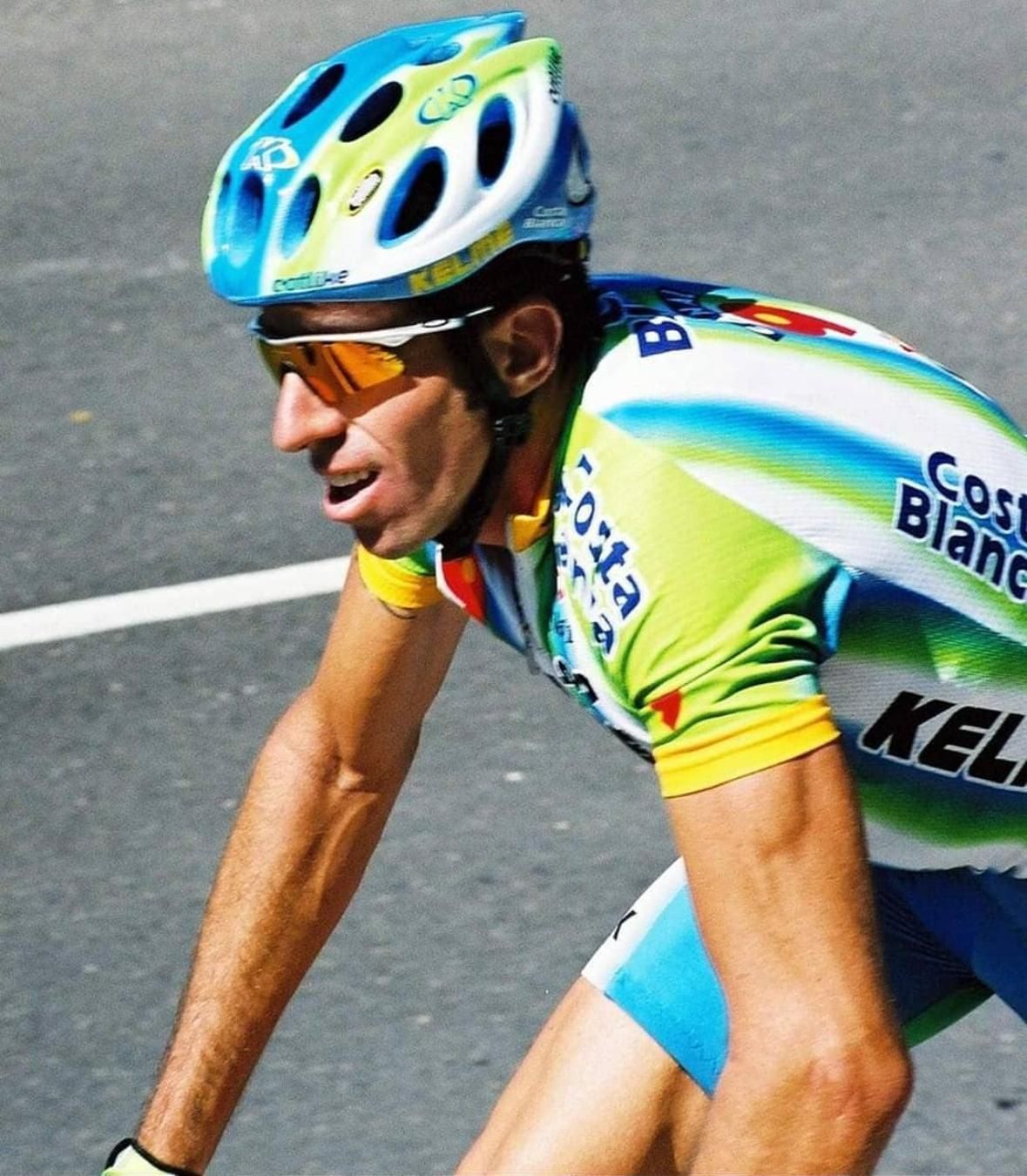 Ciclista con maillot verde y azul, casco y gafas de sol, compitiendo al aire libre.