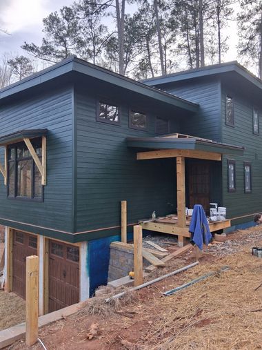 Dark green house under construction on a hillside, with unfinished lower level and exposed framing.