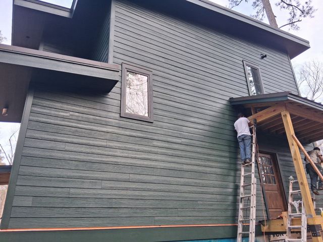 Worker on a ladder painting a dark green house exterior beside a wooden porch.