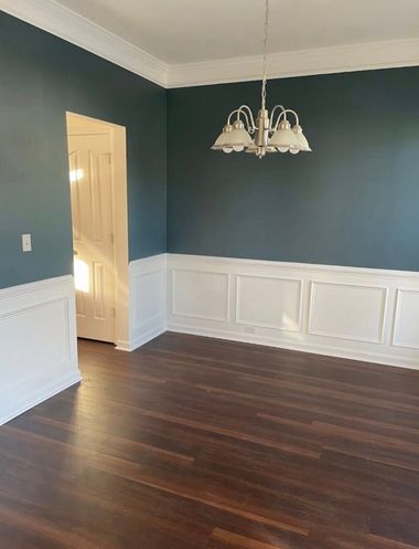Empty dining room with dark hardwood floors, teal walls, white wainscoting, and a hanging chandelier.
