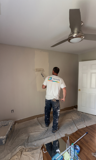 Painter working inside a room, painting a beige wall with a ceiling fan overhead and tools on the floor.