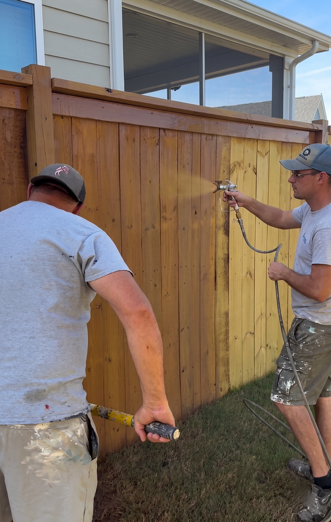 Two people spray-painting a wooden fence outdoors with a bright yellow color