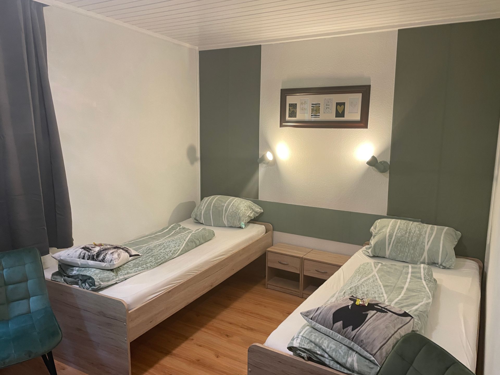 Zwei Einzelbetten in einem Zimmer mit grün-weißen Wänden, zwei Wandlampen und holzfarbenem Fußboden.