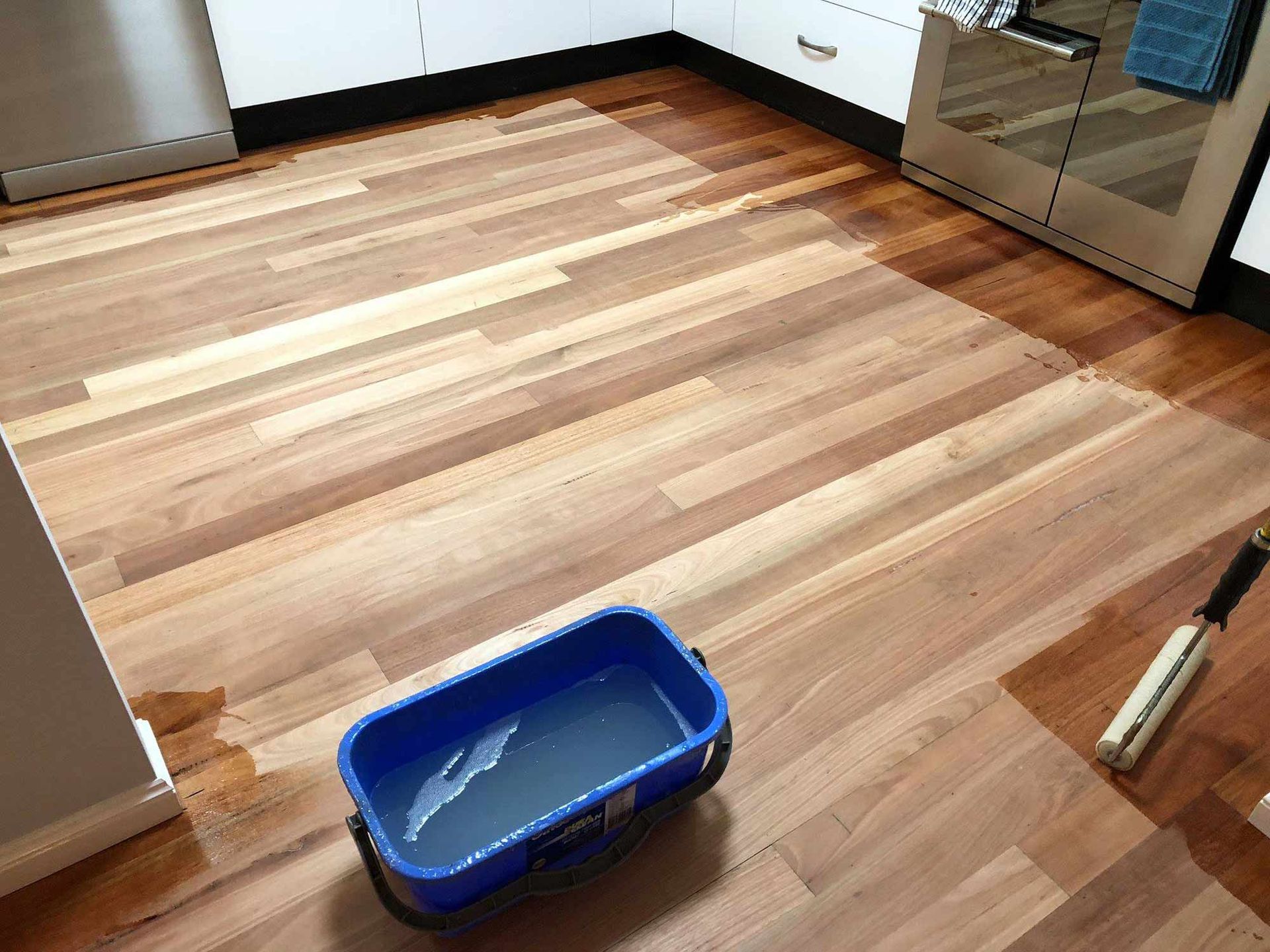 A Blue Bucket Filled With Water Is Sitting On A Wooden Floor In A Kitchen — Brett Hall Floorsanding in Port Macquarie, NSW
