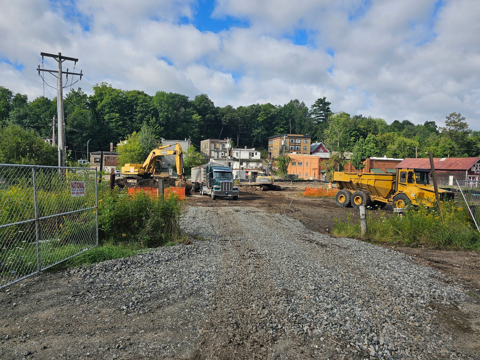 Construction site with heavy machinery, gravel road, and structures in the background under a cloudy sky.