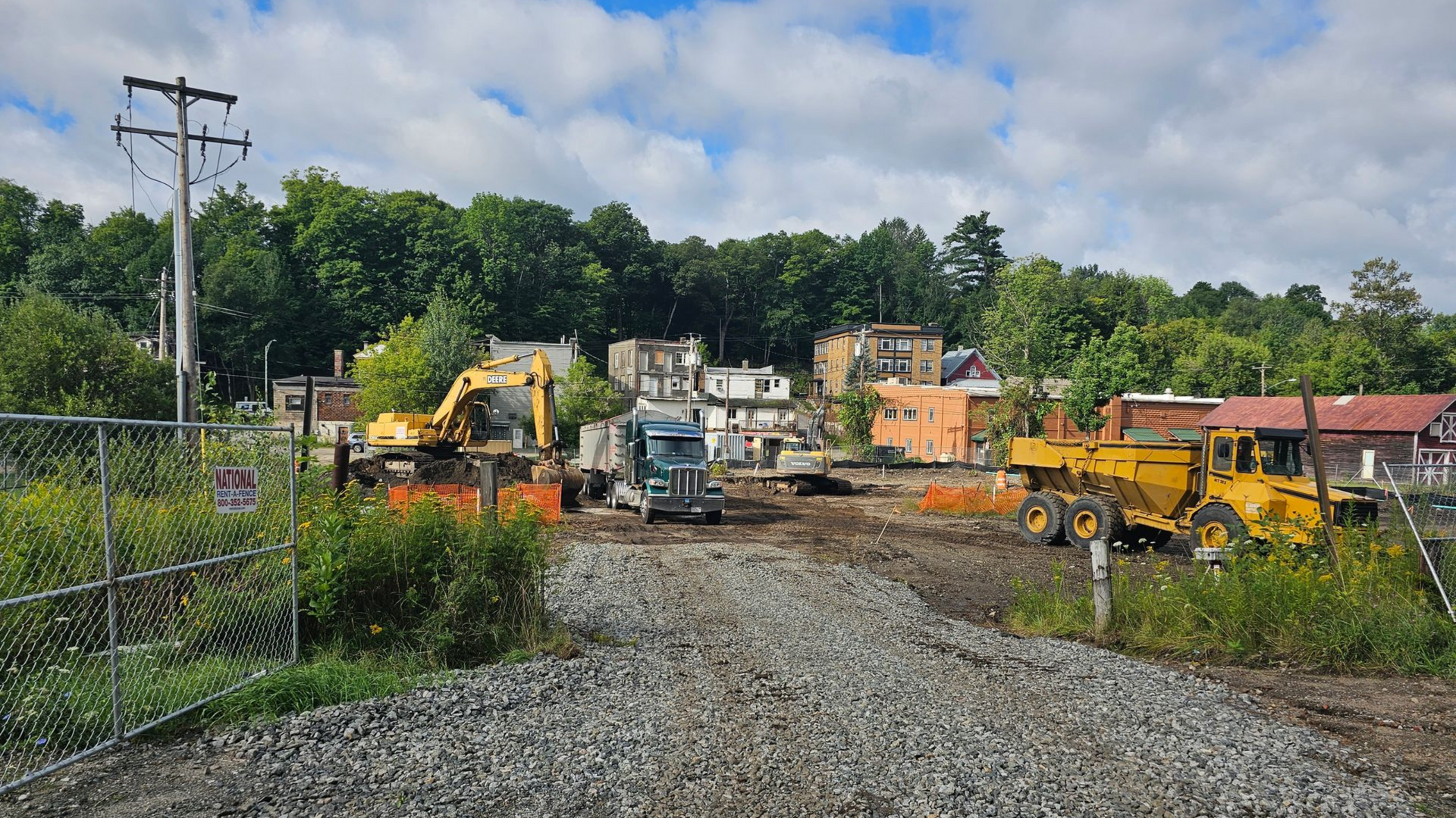 Construction site with heavy machinery: excavators, truck, and a grader. Gravel path in foreground. Buildings in background.