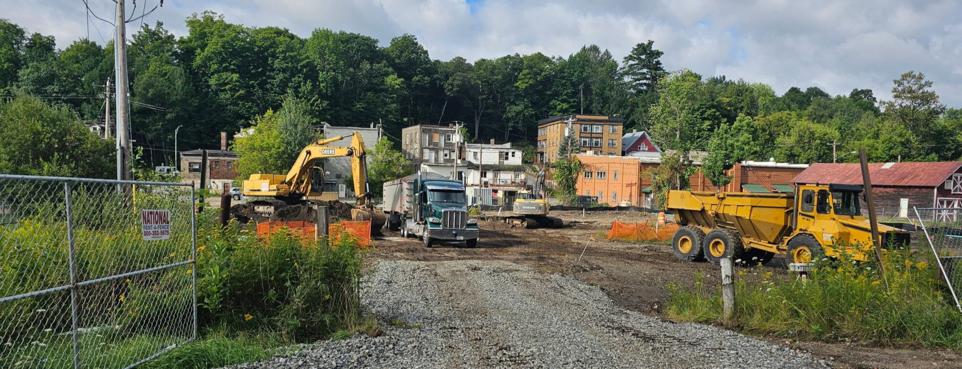 Construction site with yellow excavators, a truck, and a bulldozer clearing land near buildings and trees.