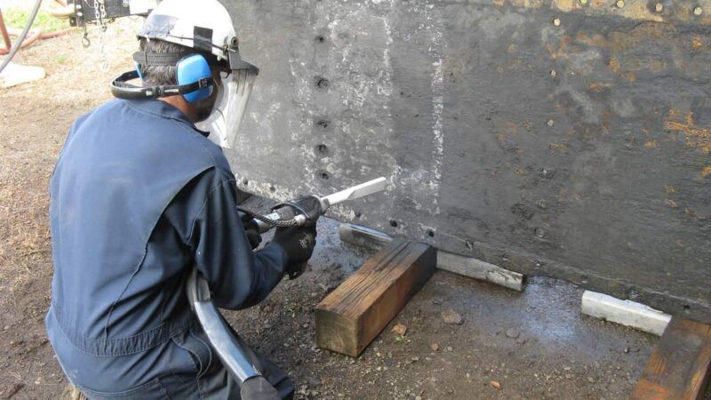 Man in safety gear sandblasting a dark, industrial metal structure outdoors.
