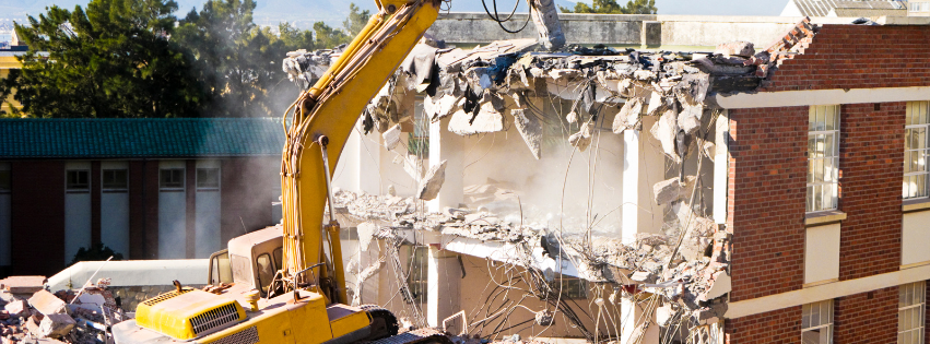 A yellow excavator is demolishing a brick building.