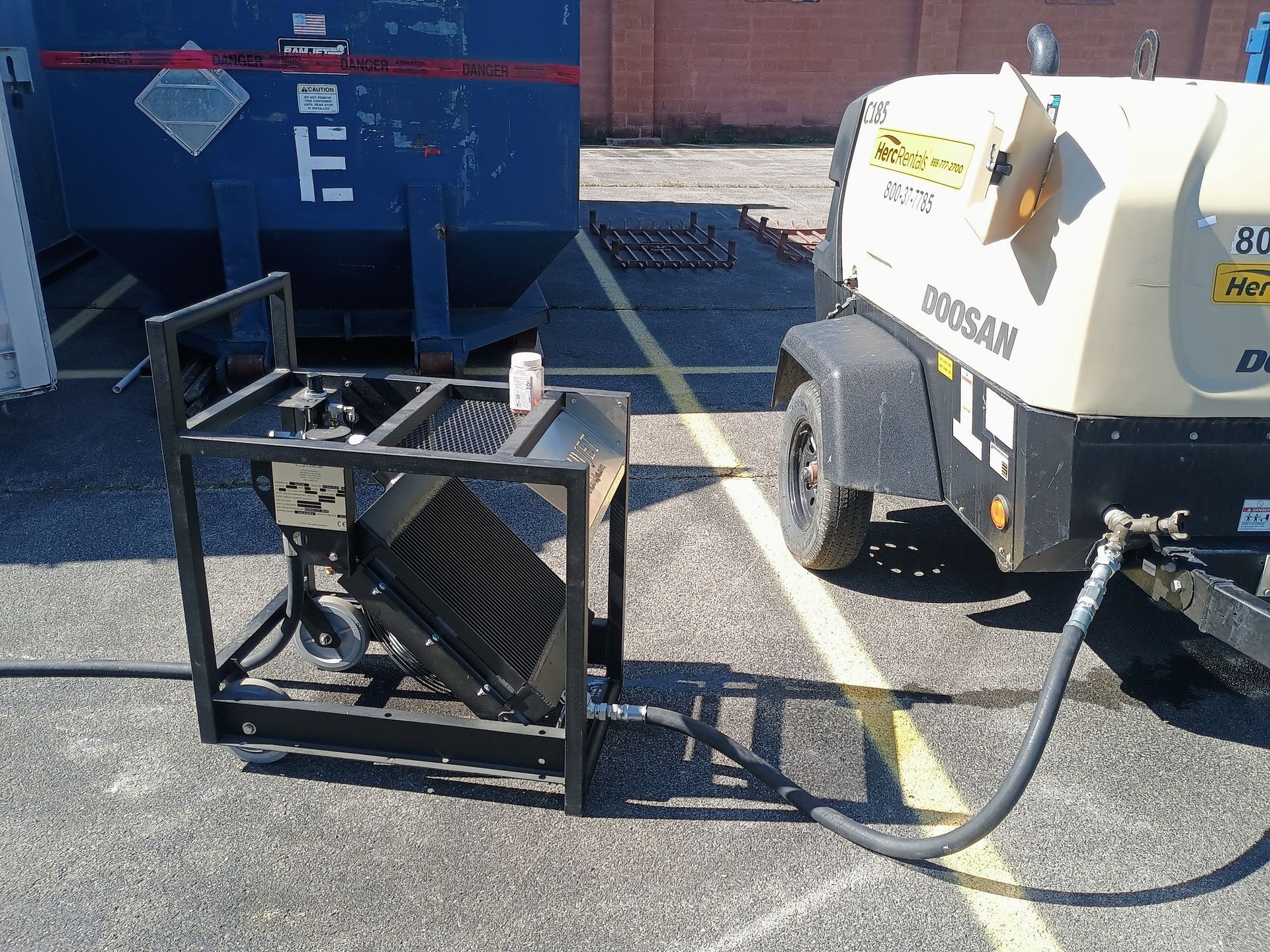 Black metal frame attached to a beige industrial machine, next to a blue dumpster on asphalt.