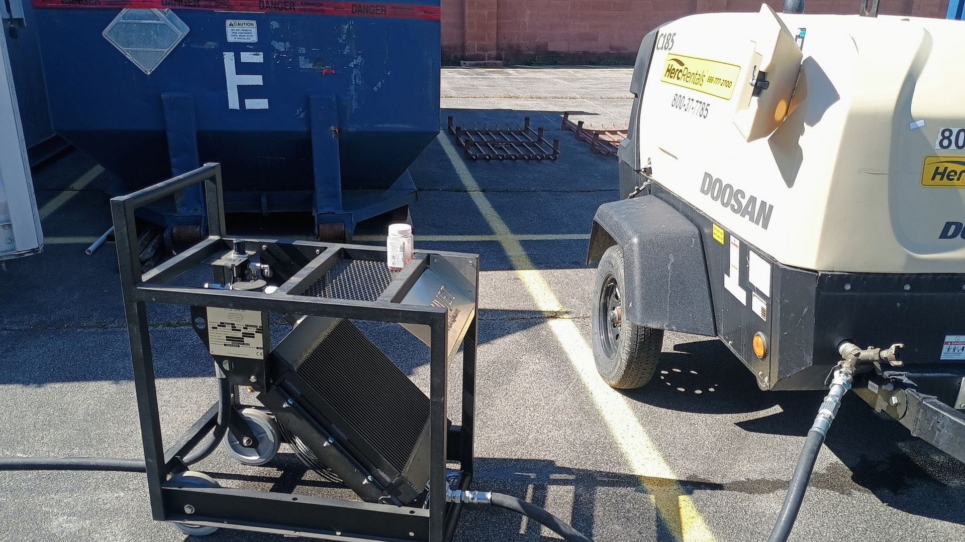Black metal equipment near a blue dumpster and an air compressor trailer parked on asphalt.