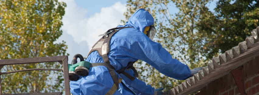 An MP Environmental employee in a blue protective suit is standing on a ladder against a roof.