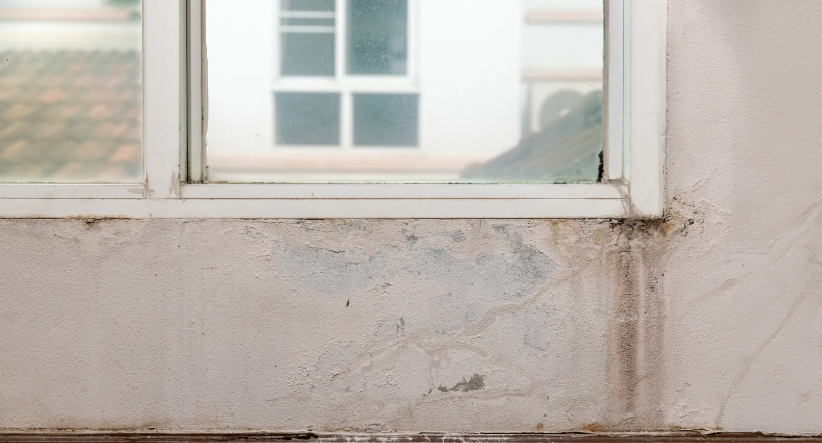 Window frame with water damage on the concrete wall. White window, light gray wall, exterior setting.