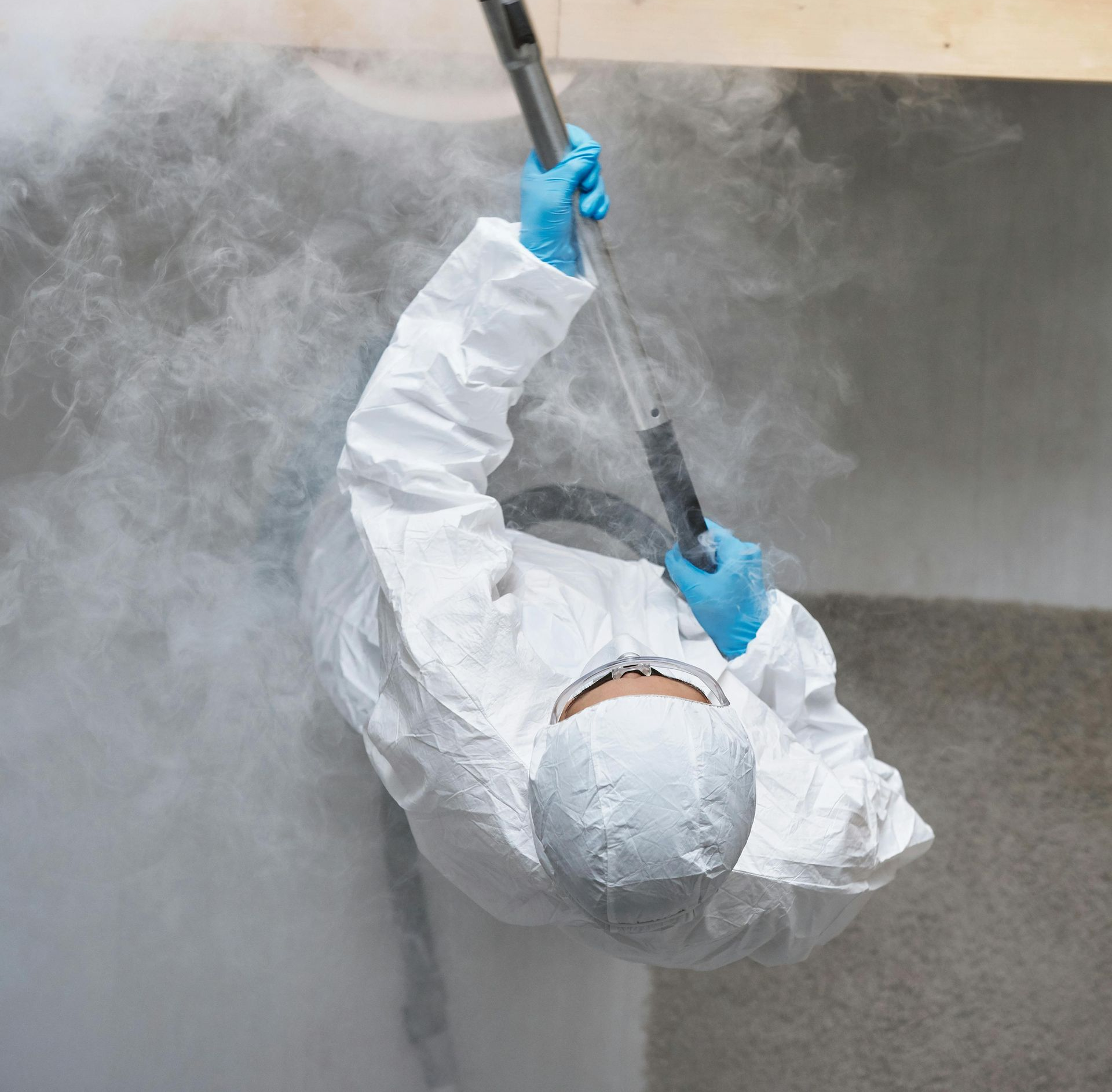 Person in protective suit spraying a mist, likely for disinfection, in an indoor setting.