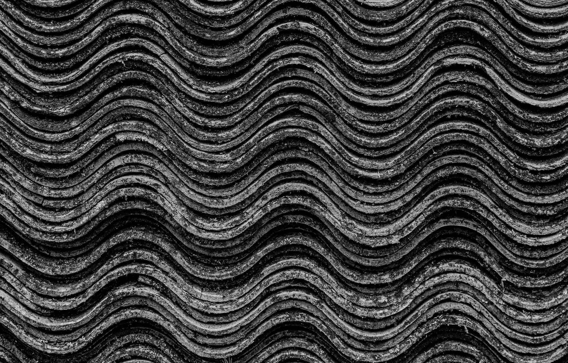 A repeating pattern of dark, weathered, corrugated asbestos roof tiles with rhythmic, wavy ridges.
