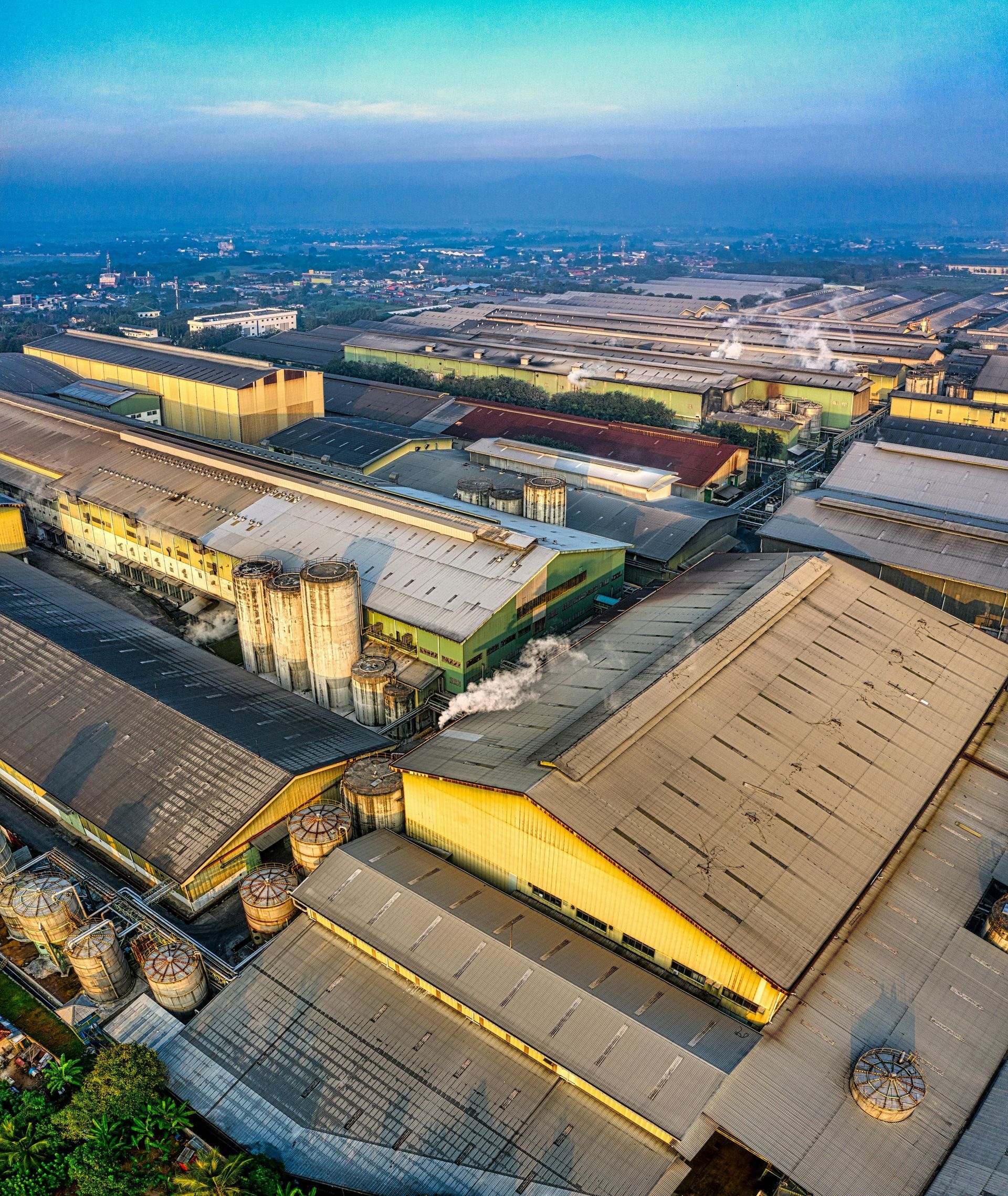 Aerial view of large industrial warehouse buildings with corrugated metal roofs and chimneys under a clear blue sky.