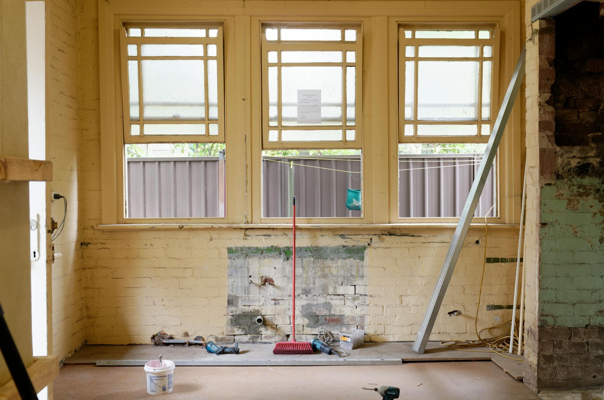 A room under renovation with three windows, an exposed brick wall section, a red broom, and supplies on the floor.