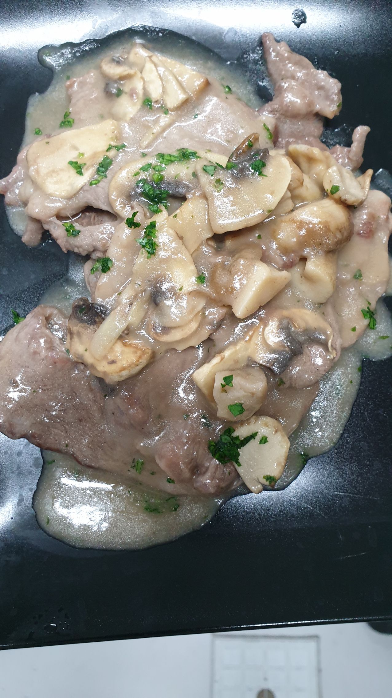 Piatto di bistecca e funghi su un piatto nero, con salsa ed erbe aromatiche tritate.