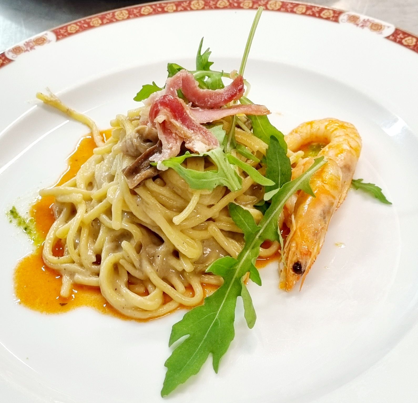 Spaghetti con gamberi, prosciutto e rucola su un piatto bianco con bordo rosso.
