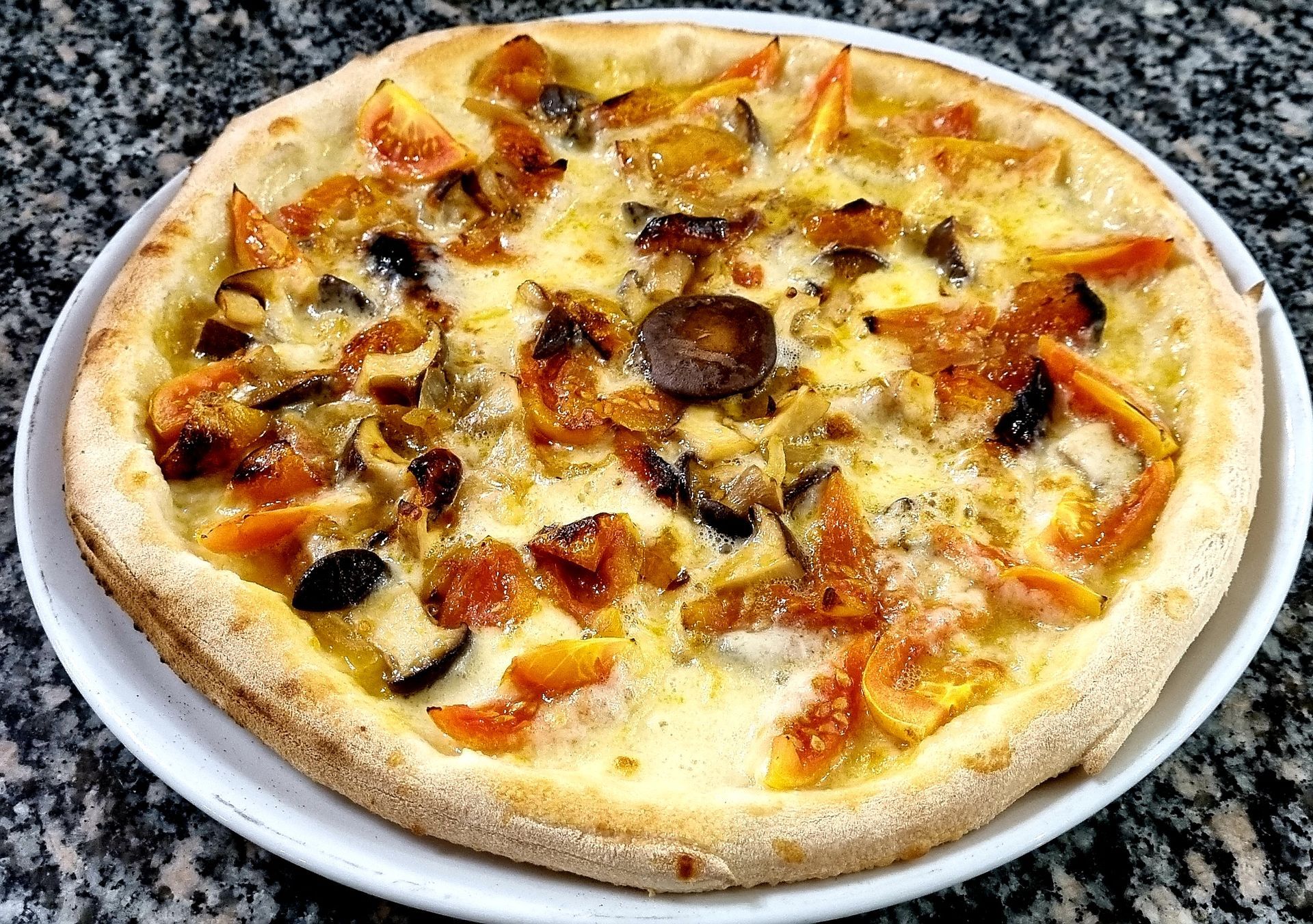 Pizza su un piatto bianco, con condimenti tra cui pomodori, funghi e olive, su una superficie di marmo.