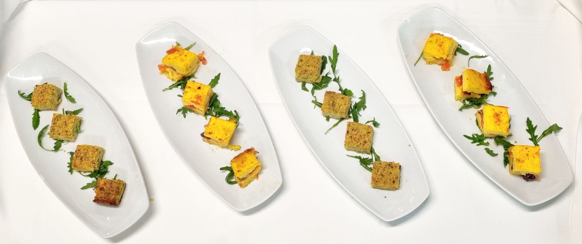Quattro piatti bianchi con antipasti a cubetti, guarnizione di rucola e sfondo bianco.