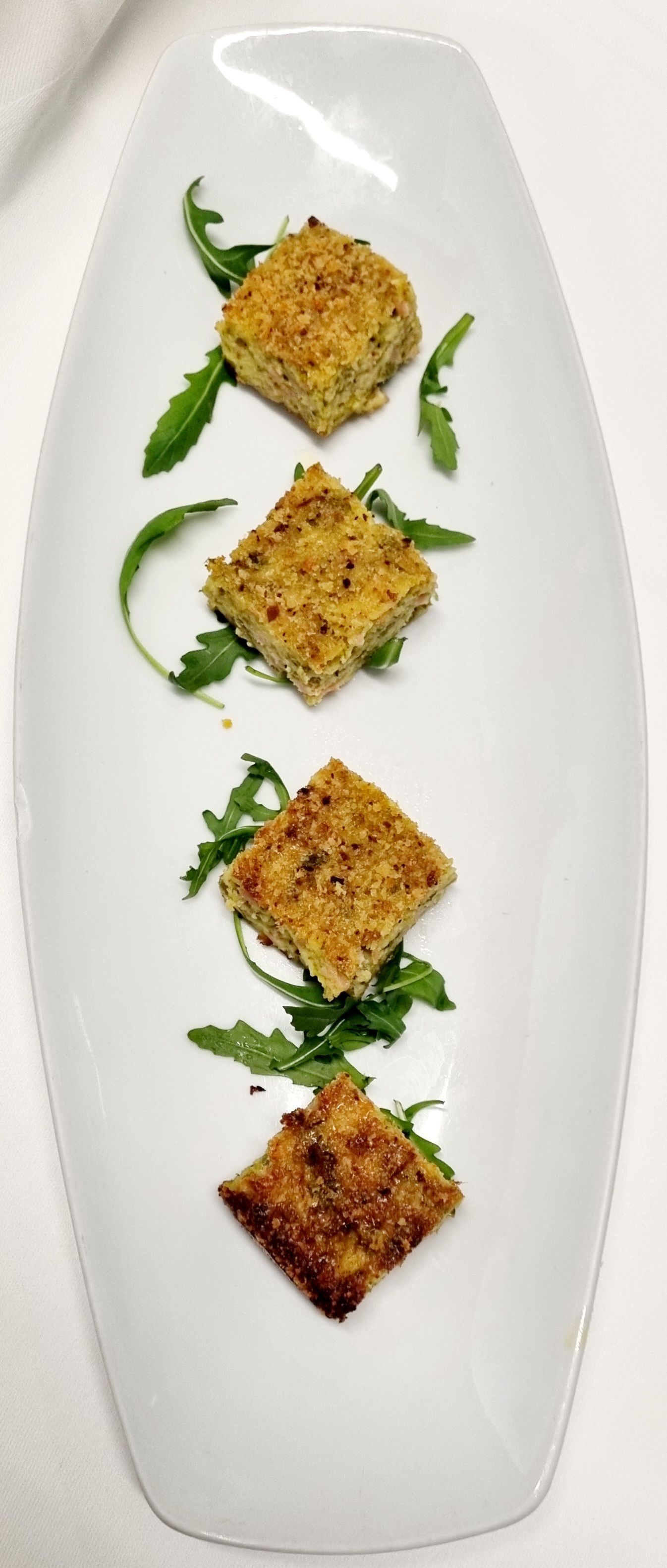 Quattro antipasti quadrati impanati con rucola su un piatto ovale bianco.