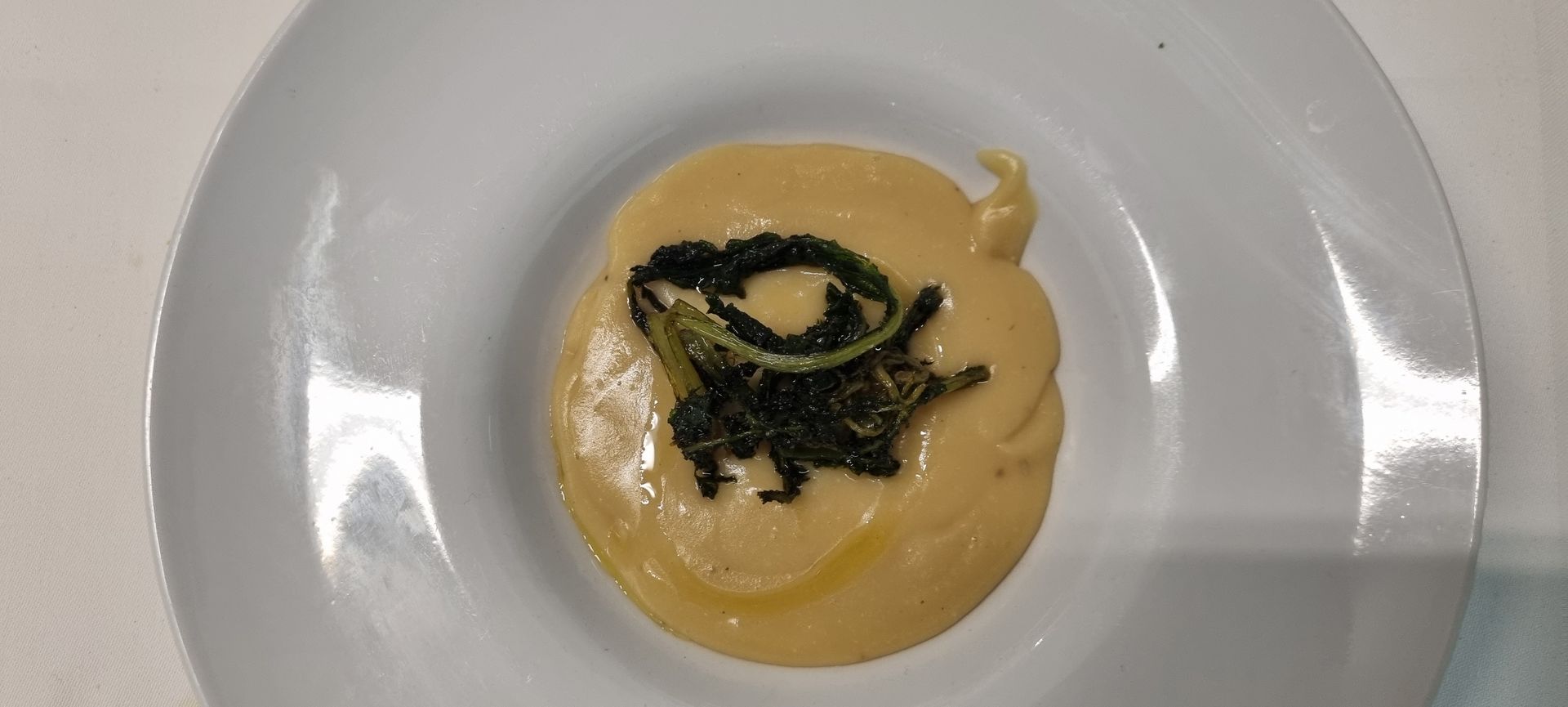 Cibo cremoso di colore giallo guarnito con verdure a foglia verde scuro su un piatto bianco.