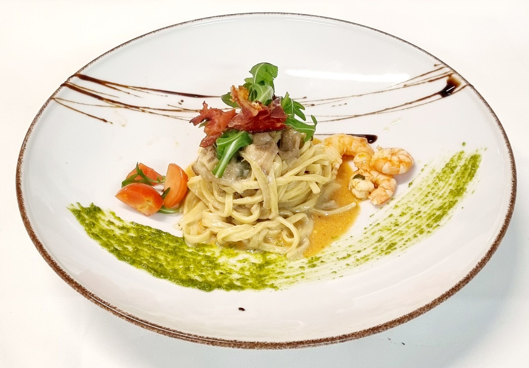 Piatto di pasta con pesto, pomodori, gamberi e prosciutto su un piatto bianco.