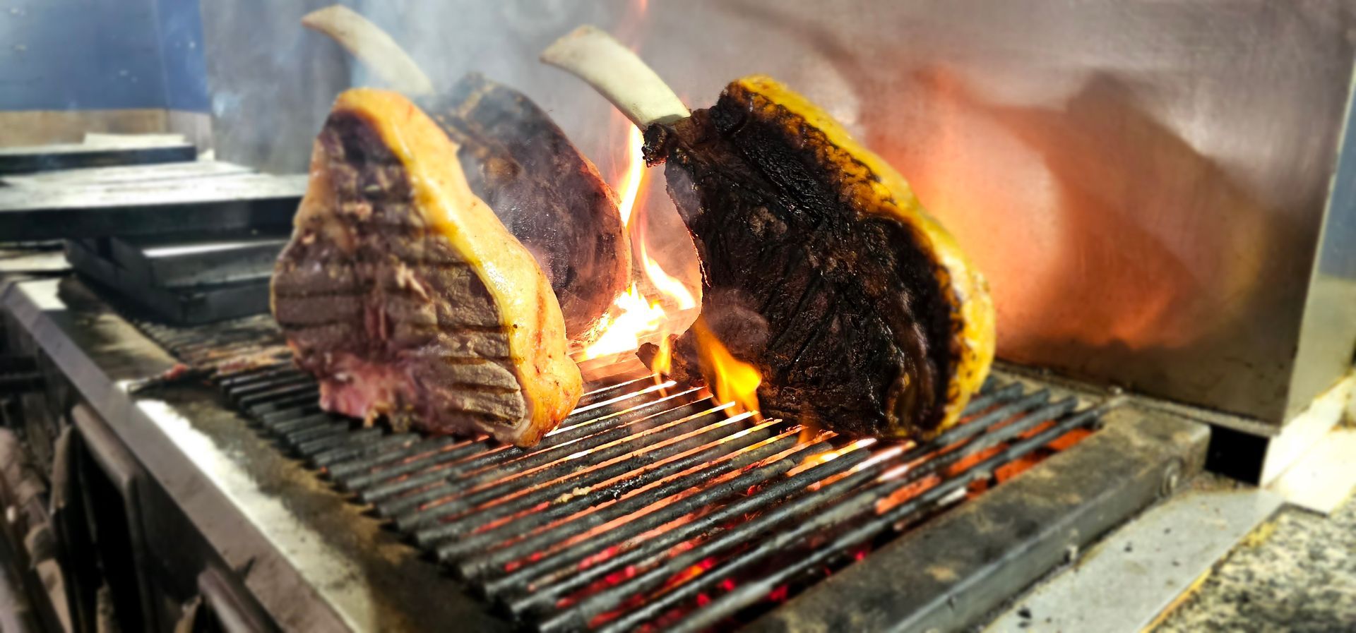 Tre grandi bistecche cotte sulla griglia metallica calda, con le fiamme visibili.