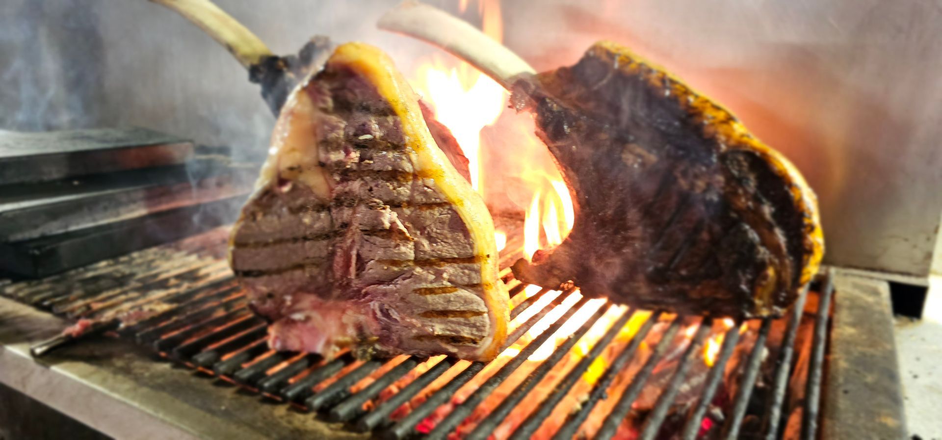 Grandi bistecche cotte su una griglia, con le fiamme visibili, in una cucina professionale.