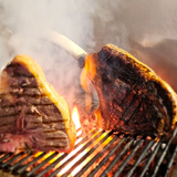 Due grandi bistecche cotte alla griglia su una fiamma viva, su una griglia metallica, producendo fumo.
