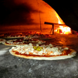 Pizze condite in un forno a legna, con fiamme sullo sfondo, pronte per essere cotte.