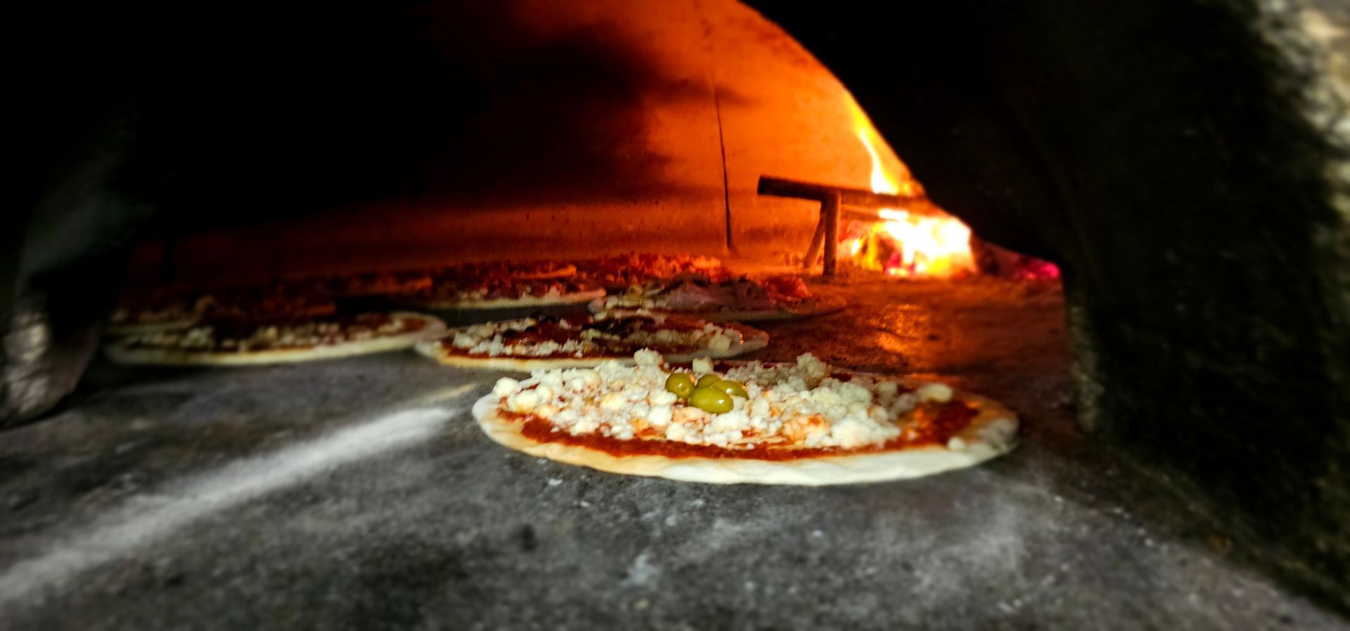 Pizze cotte nel forno a legna, vicino a un fuoco scoppiettante.