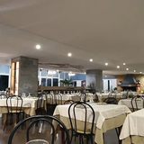 Interno di un ristorante con tavoli apparecchiati per la cena. Tovaglie beige, sedie nere e illuminazione dall'alto.